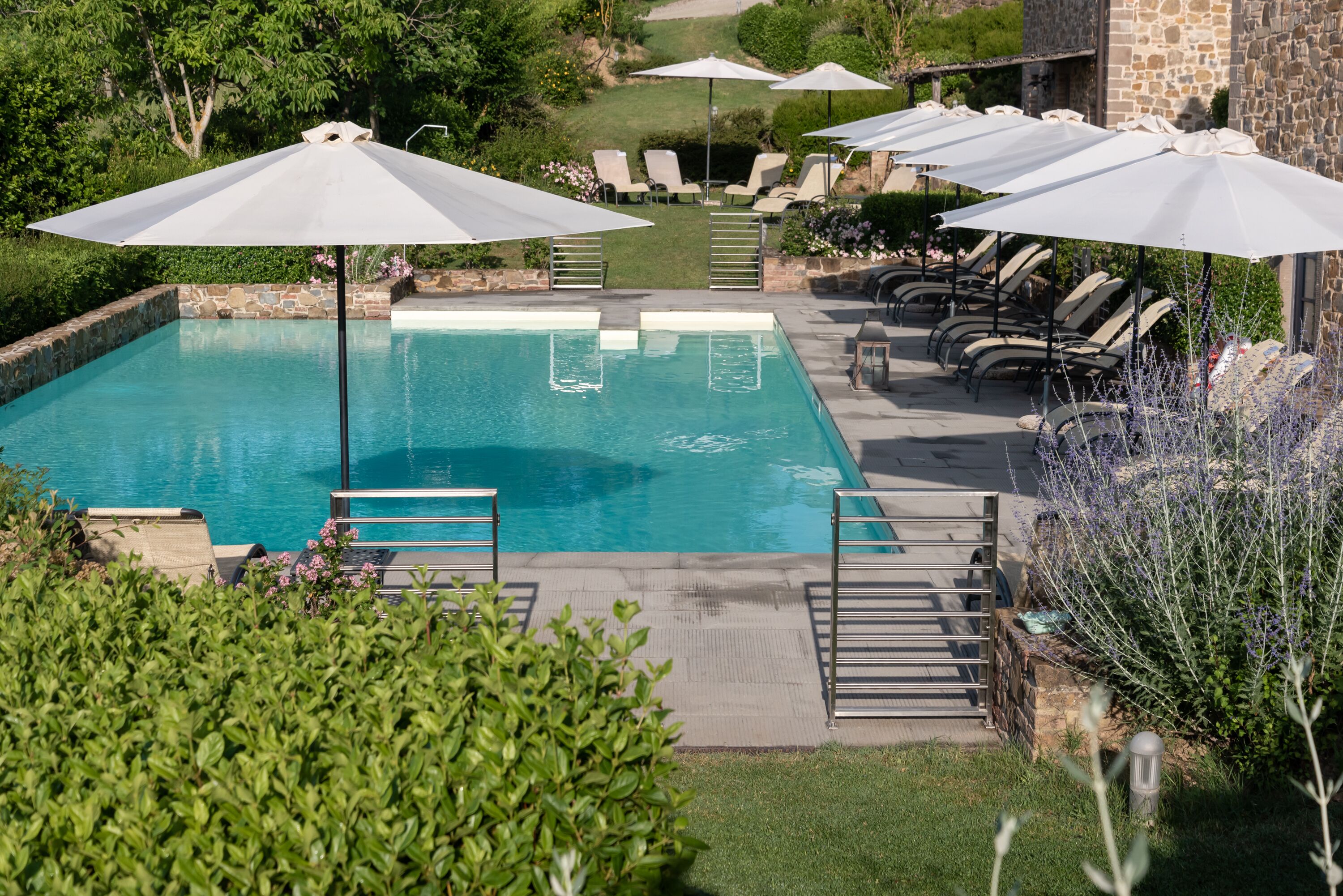 A pool area with lounge chairs and umbrellas, surrounded by greenery and a stone building.