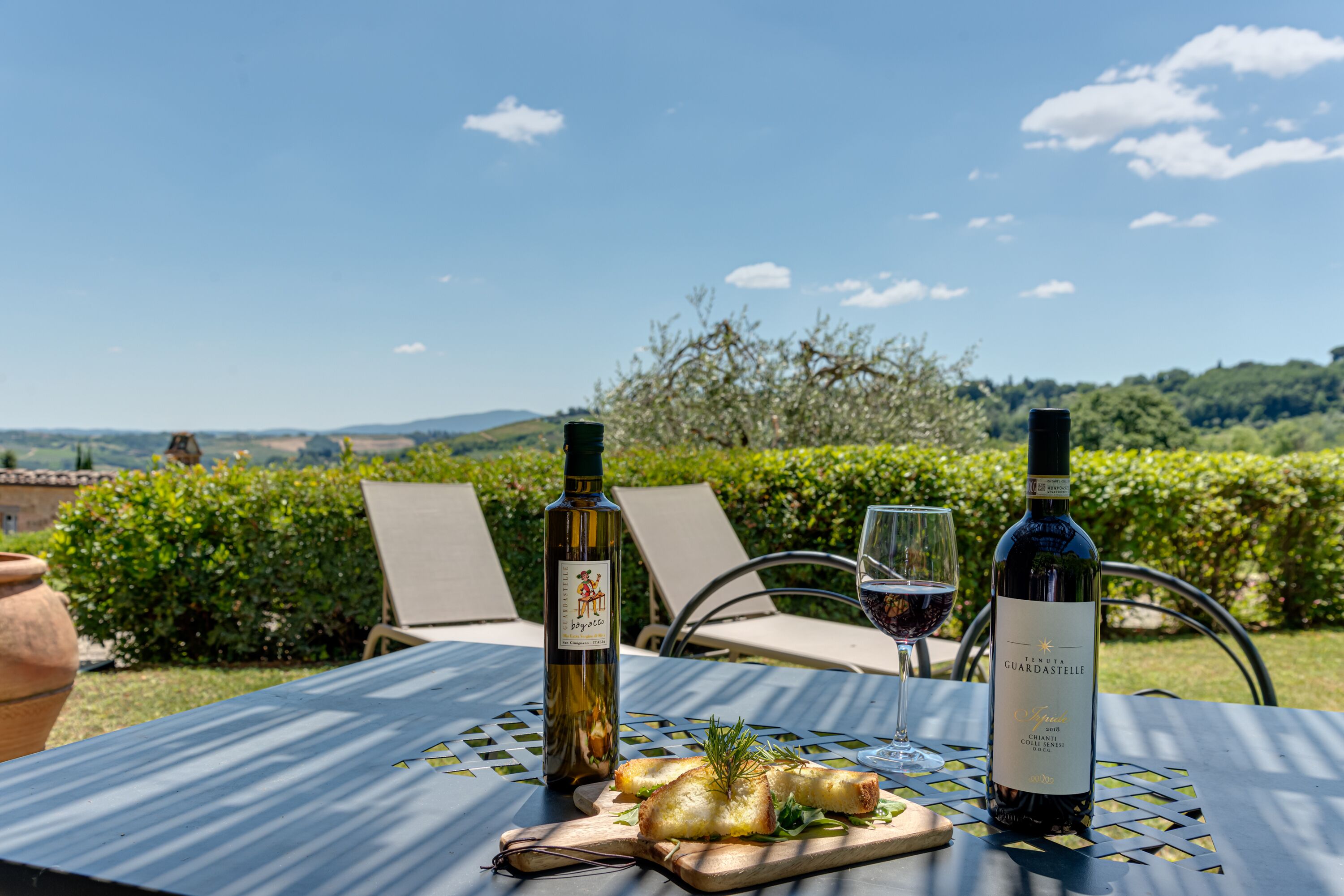 Una mesa con dos botellas de vino y una copa de vino tinto, una tabla de madera con comida, y una vista panorámica de fondo.