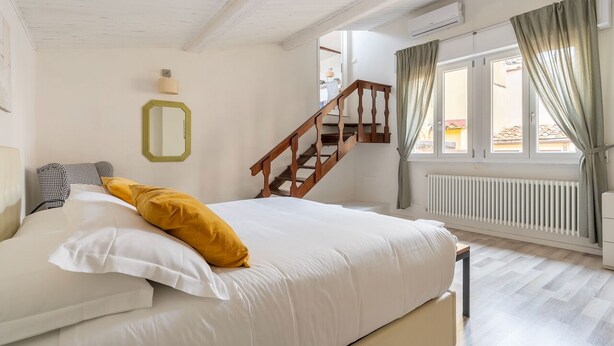 A bedroom with a bed, a wooden staircase, a window with curtains, and a radiator. - House on the Roof - Florence Dolce Vita (Firenze)