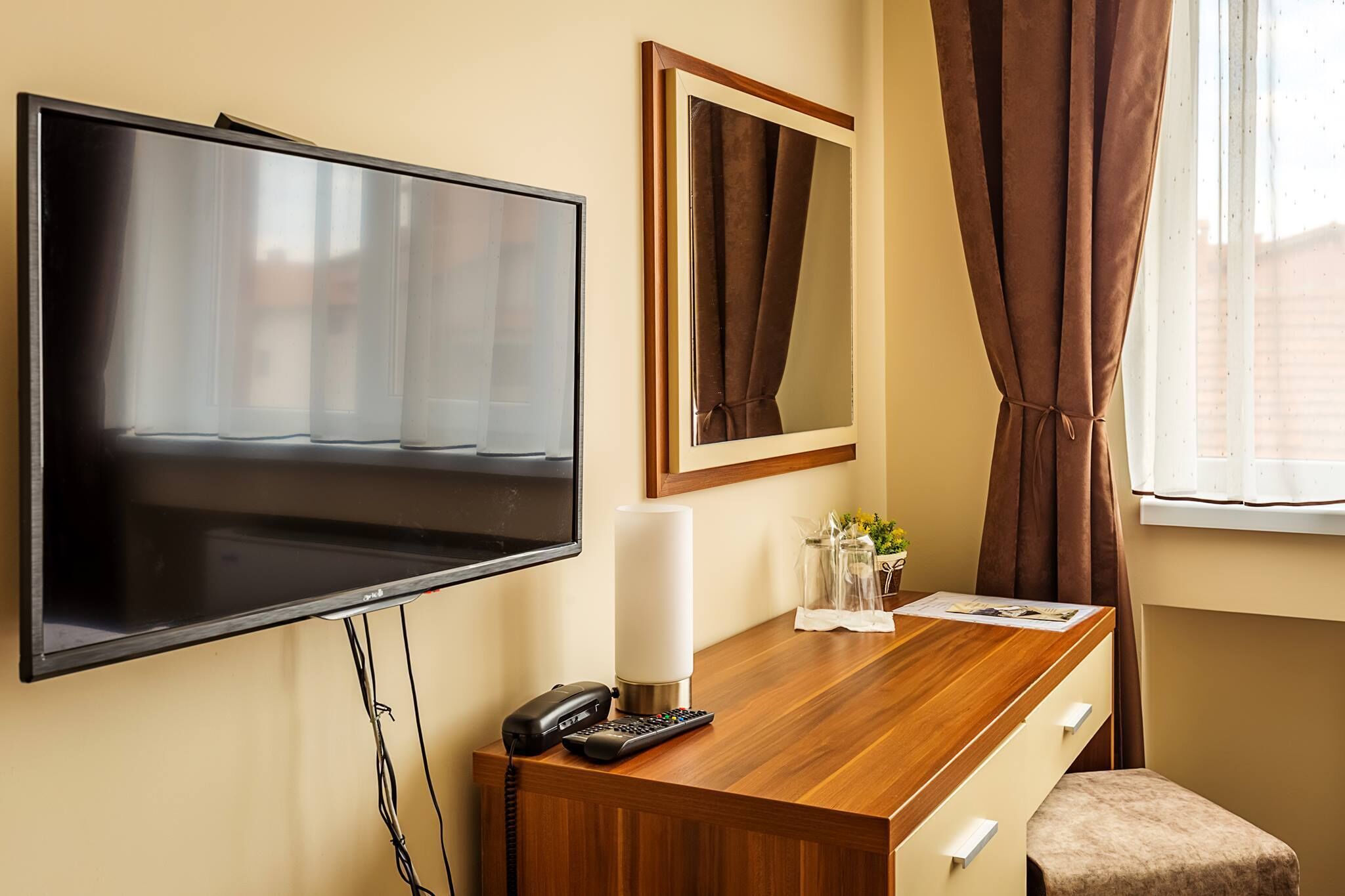 Habitación de hotel con televisor de pantalla plana, escritorio con teléfono y control remoto, espejo, florero con flores y ventana con cortinas.