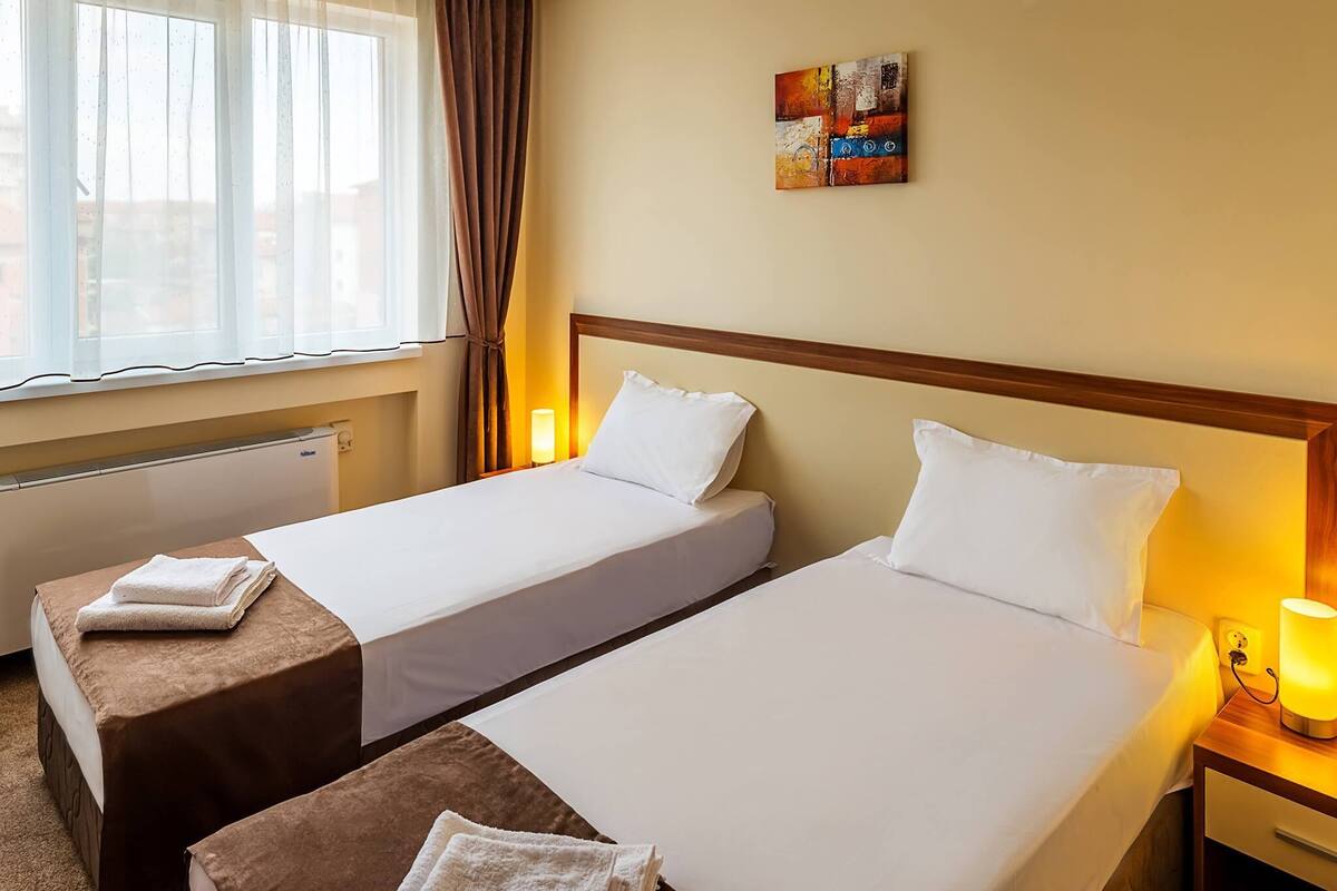 A hotel room with two single beds, a radiator, a window with curtains, a wall-mounted abstract painting, and a bedside lamp.