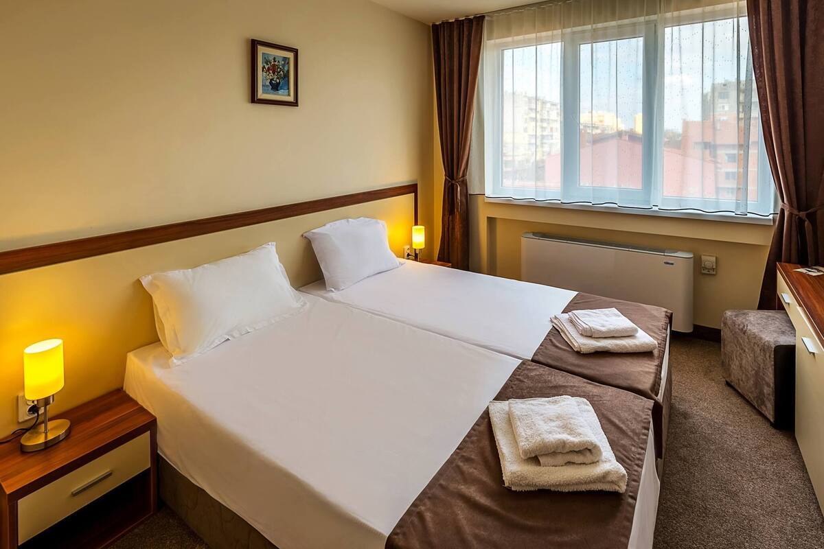A hotel room with two beds, a nightstand with a lamp, a window with curtains, and a small table with towels.