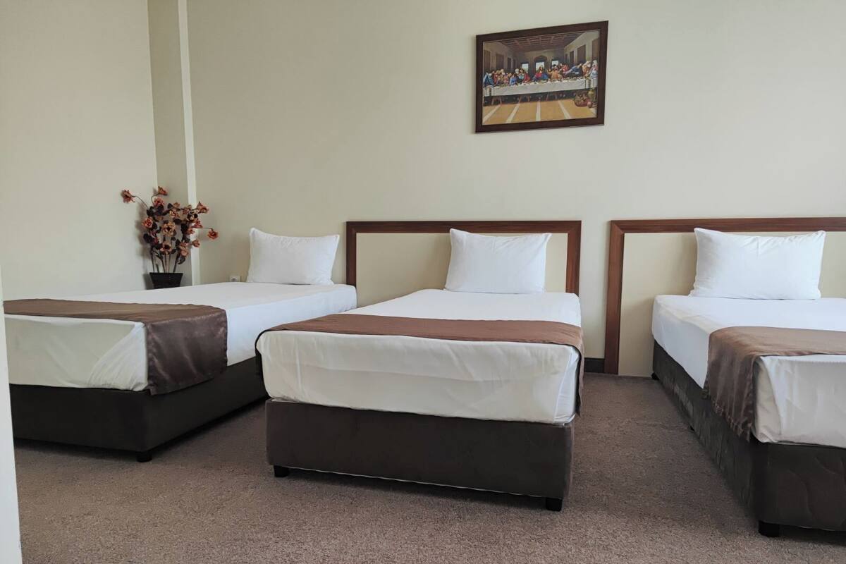 A hotel room with three beds, a painting on the wall, and a vase with dried flowers.