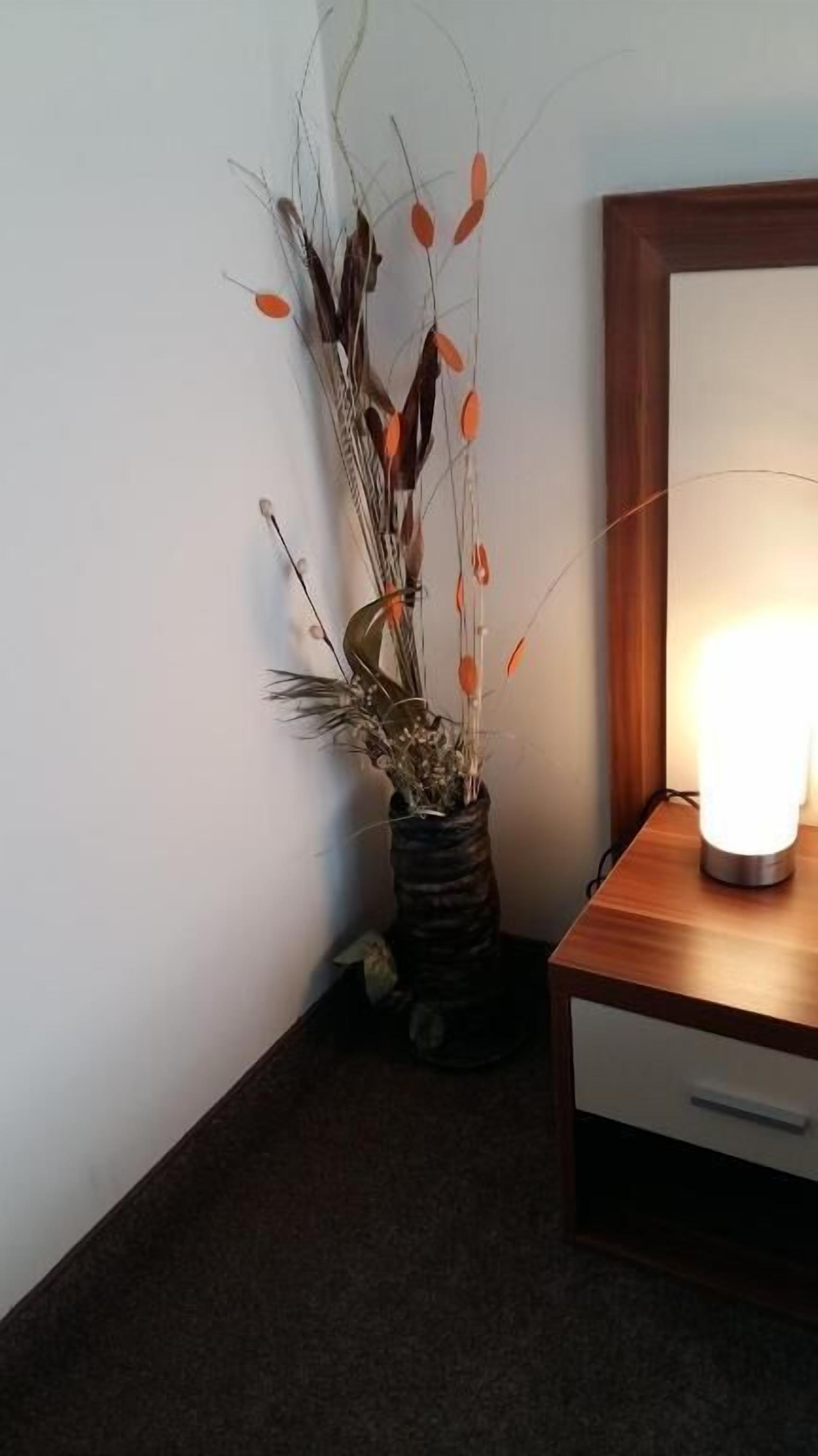 Rincón de una habitación con una mesita de noche de madera, una lámpara y un florero con plantas secas.