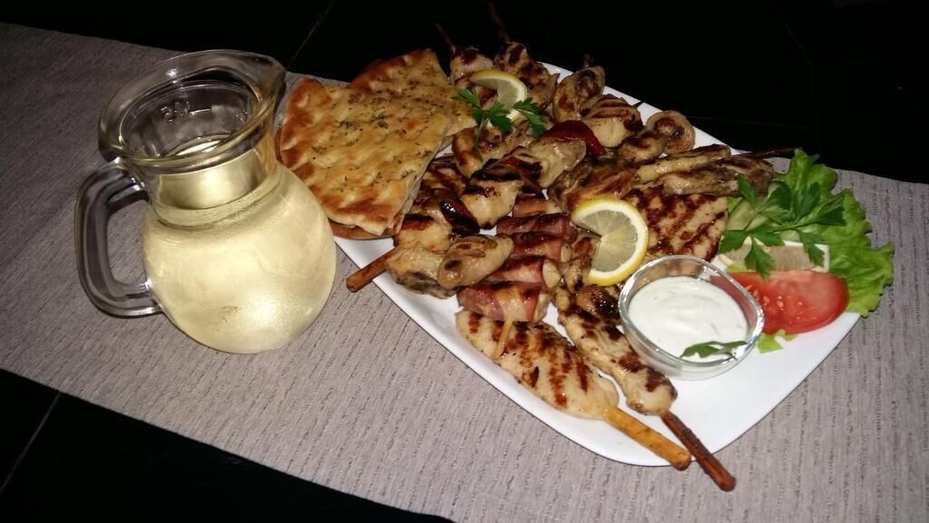 A plate of grilled chicken skewers, a slice of flatbread, a glass pitcher, and a small bowl of sauce, accompanied by fresh vegetables.