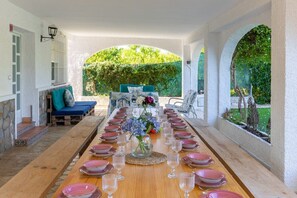 A long wooden dining table set for a meal with pink plates and clear glasses, surrounded by white walls and a view of greenery.