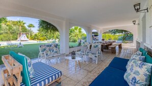 Área de estar coberta ao ar livre com vista para a piscina, móveis de madeira e mesa de jantar para quatro pessoas.