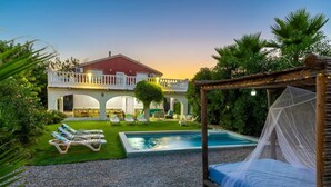 Uma área de piscina com espreguiçadeiras, uma cama com dossel e um edifício com varanda.