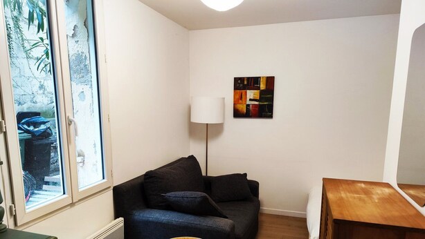 A compact living space with a dark sofa, a small round table, a radiator, and a window with a view of greenery. - Charming apartment downtown Bordeaux (Bordeaux)