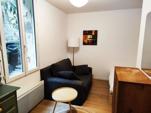 Un espace de vie compact avec un canapé foncé, une petite table ronde, un radiateur et une fenêtre donnant sur un paysage verdoyant.