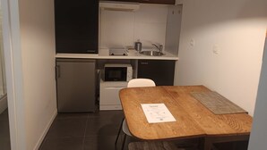 Une cuisine compacte équipée d’un four à micro-ondes encastré, d’un petit réfrigérateur et d’un évier. On y trouve une table en bois, un paillasson et un document posé dessus.