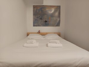Een netjes opgemaakt bed met witte lakens en twee opgerolde handdoeken ervoor, een houten hoofdbord en een landschapsschilderij erboven.