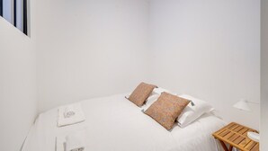 Une petite chambre blanche, avec un lit simple, une table de chevet en bois et une petite table sur laquelle se trouve un lavabo blanc.