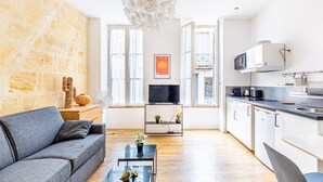 Ein modernes Apartment mit einer Wand aus Stein, einem grauen Sofa, einem Couchtisch, einem Fernseher und einer Küche mit weißen Schränken und einer schwarzen Arbeitsplatte.