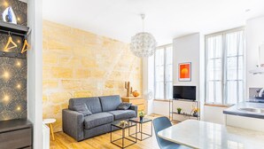 Ein modernes Wohnzimmer mit Steinwand, einem grauen Sofa, einem Esstisch mit Stühlen, einem Fernseher und einer Küchenzeile mit Spüle und Fenster.
