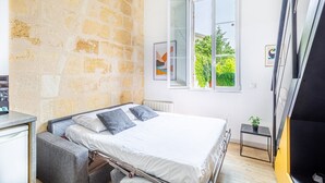 A bedroom with a stone wall, a bed with white bedding, a window with a view of greenery, and a small bedside table with a plant.