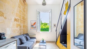 A compact living space with a stone wall, a gray sofa, a small table, a yellow cabinet, and a large window offering a view of greenery.