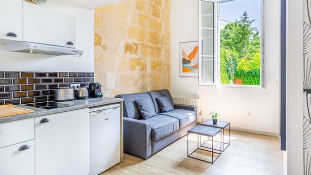 A modern kitchen with white cabinets, a stone accent wall, a grey sofa, and a window with a view of greenery.