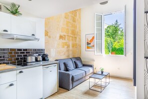 A modern kitchen with white cabinets, a stone accent wall, a grey sofa, and a window with a view of greenery. - Cosy duplex apartment Bordeaux center (Bordeaux)