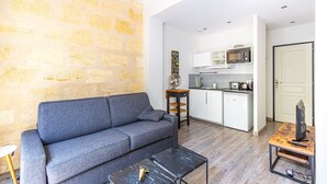 A compact living space with a grey sofa, a small coffee table, a fan, and a kitchenette with white cabinets and a microwave.