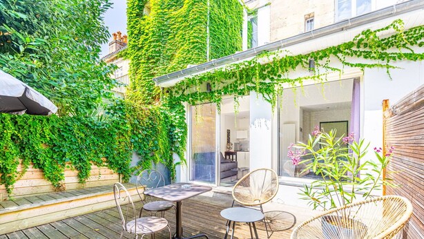 A wooden deck with a table and chairs, surrounded by lush greenery and a building with a glass door.