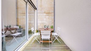 A small patio with a wooden table and white chairs, surrounded by stone walls and a glass door.