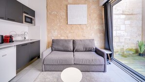 A compact living space with a kitchenette, a grey sofa, a white table, and a view of the outside through a large window.