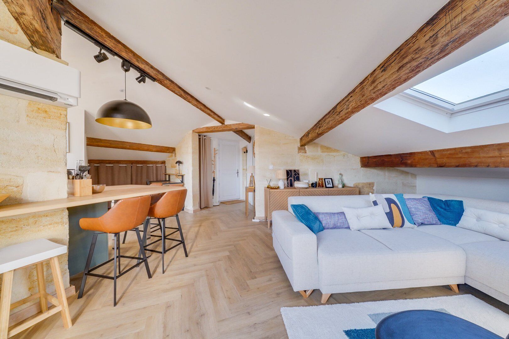 Ein großzügiger Wohnbereich mit hoher Decke, freiliegenden Holzbalken, einem weißen Sofa mit bunten Kissen, einer Holztresore und Barhocker sowie einem Oberlicht.