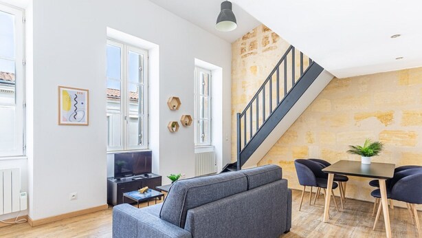 A modern living room with a grey sofa, a dining table with chairs, a TV, and a staircase against a textured wall. - Beautiful apartment with Parking at Bordeaux (Bordeaux)