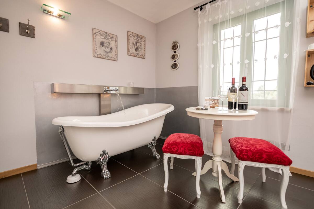 A bathroom with a clawfoot bathtub, a round table with two red cushioned stools, and a window with curtains.