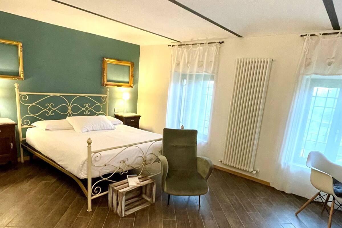 A bedroom with a bed, two chairs, a nightstand, a bedside table, and a window with curtains.