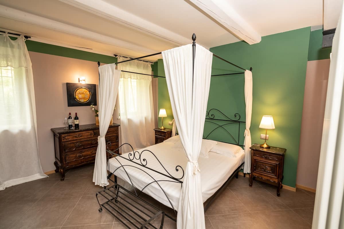 A four-poster bed with white linens, a wooden nightstand with a lamp, and a dresser with bottles and a vase.