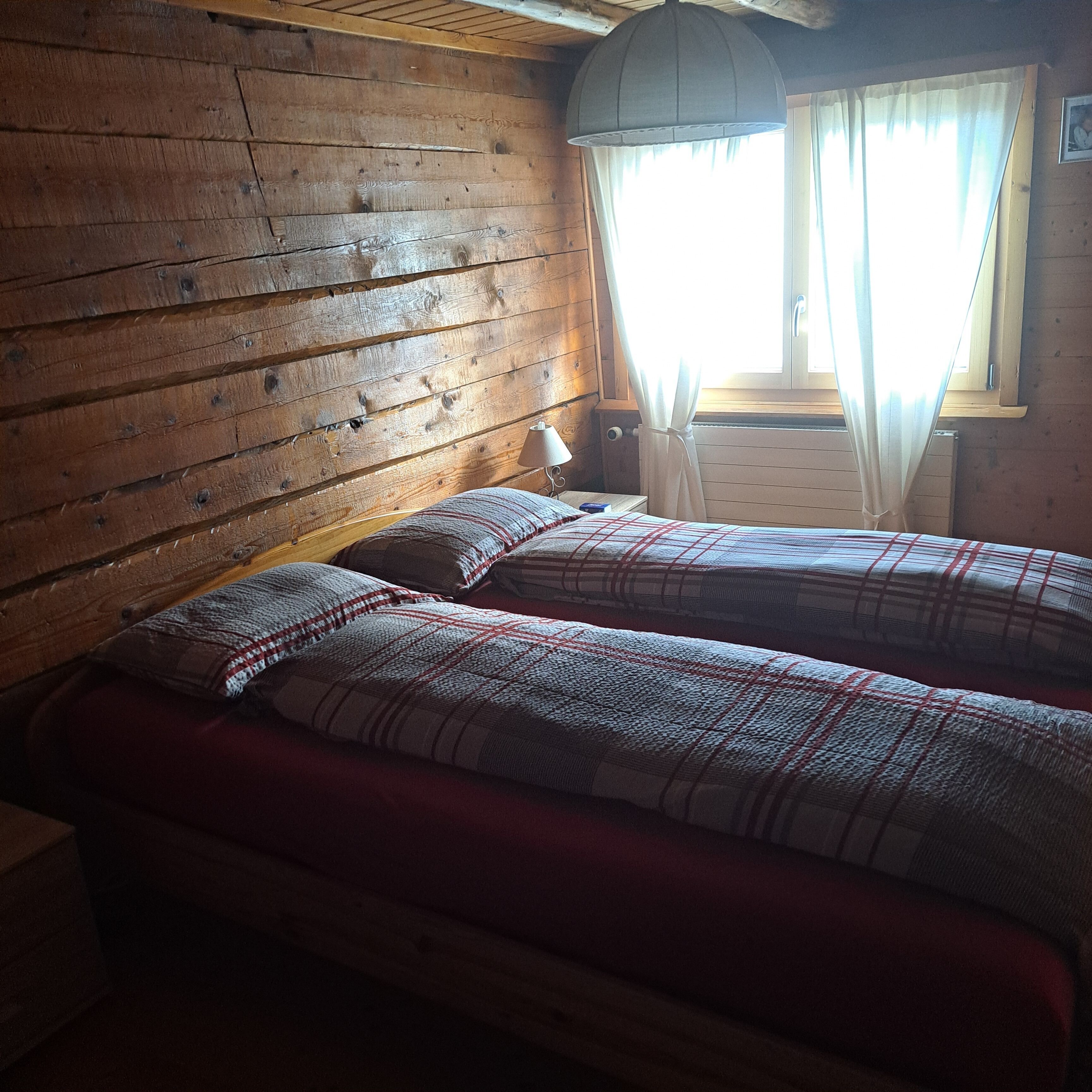Ein Zimmer in einer Holzhütte mit zwei Betten, einem Fenster mit Vorhängen und einer Lampe auf einem Nachttisch.