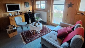 Interior acolhedor de cabana com teto de madeira, televisão, sofá, uma pequena mesa com uma planta e janela decorada com estrelas.