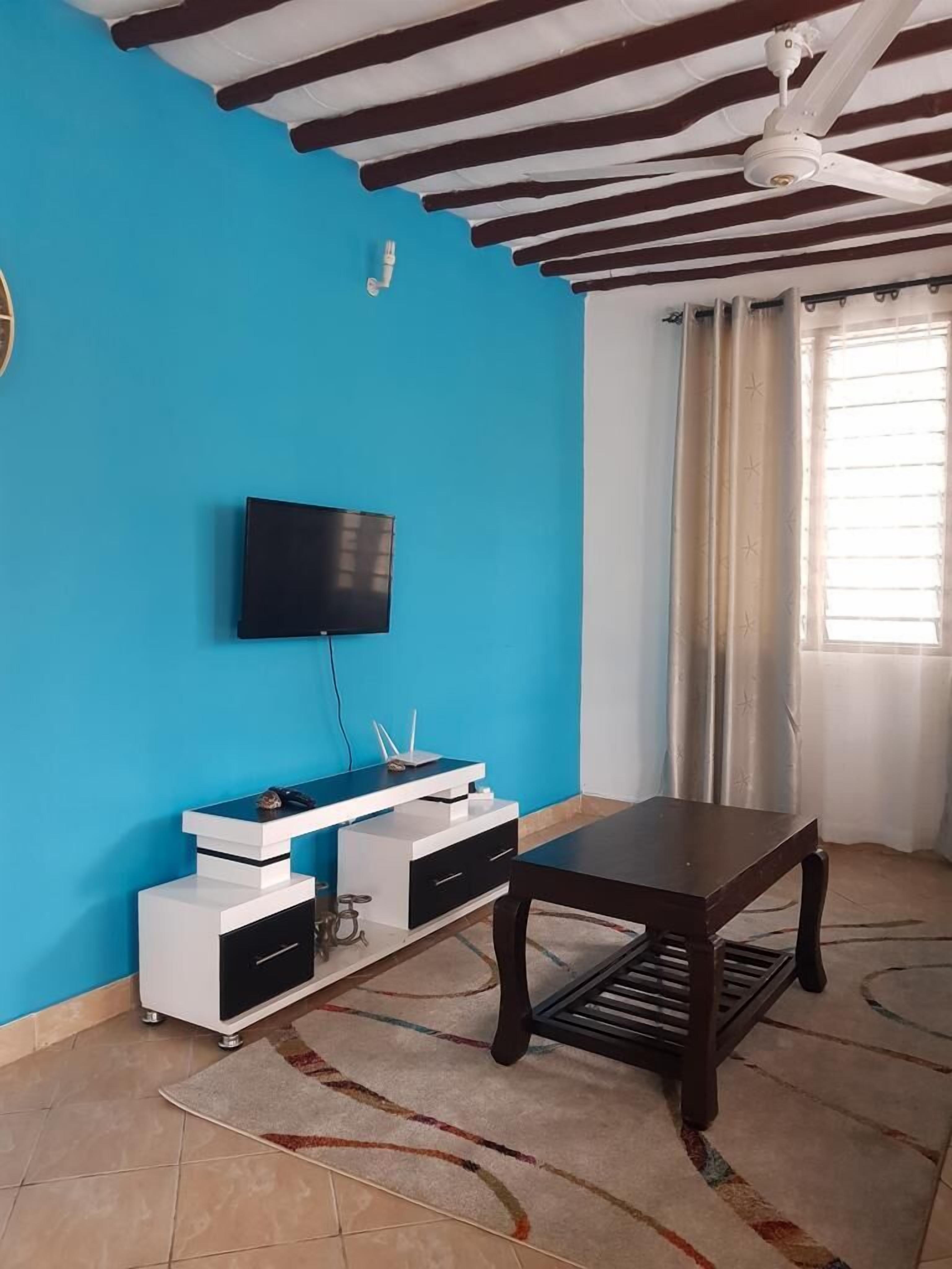 Une pièce avec un mur bleu, un téléviseur à écran plat fixé au mur, une table en bois et un ventilateur de plafond.