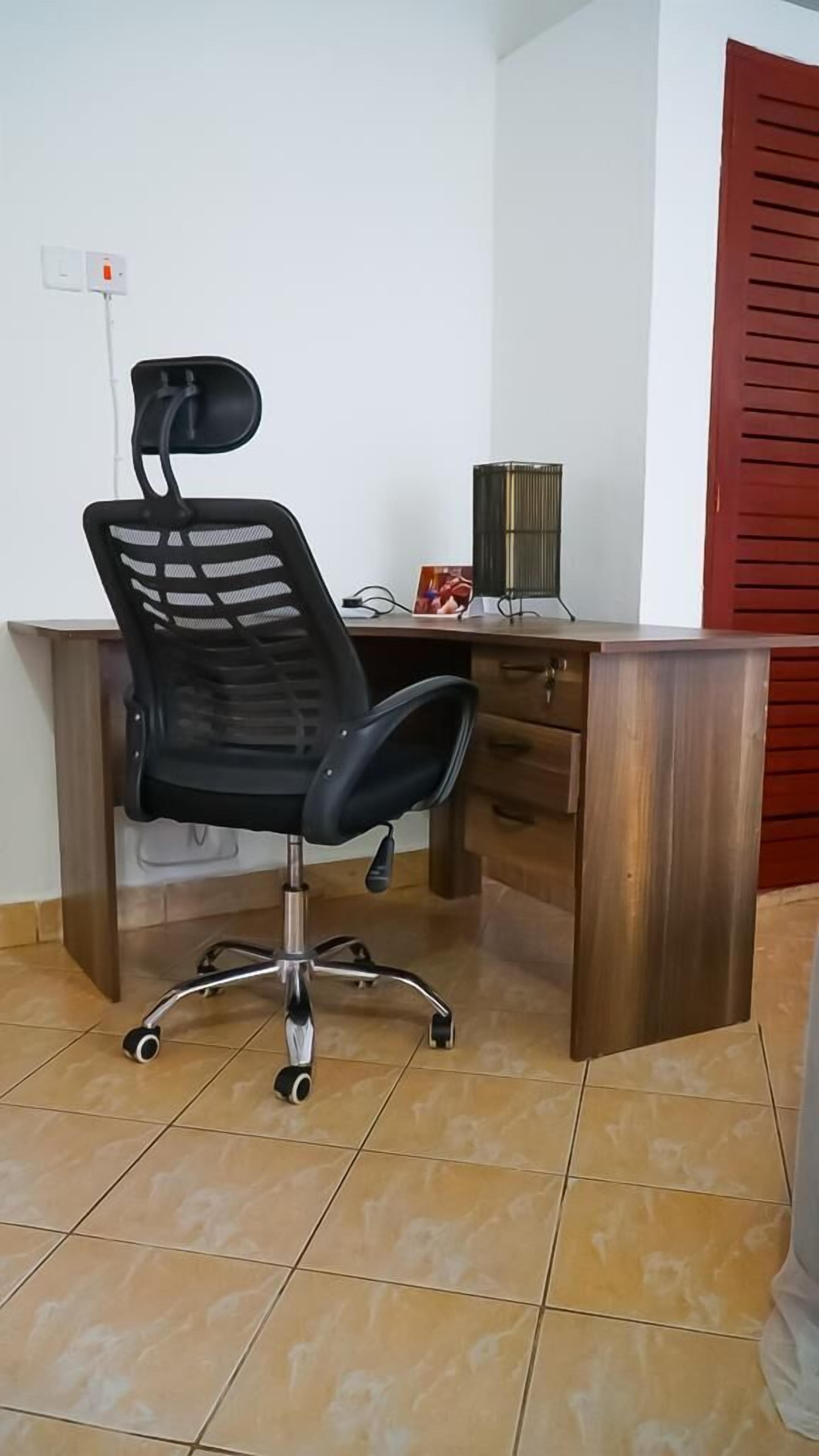 A corner office with a black office chair, a wooden desk, a red door, and a tiled floor.