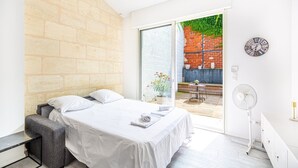 Un dormitorio con una cama, una mesita, una silla, un ventilador y vistas a un patio exterior con pared de ladrillo y un reloj.
