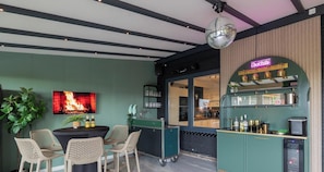 Un espace bar agrĂ©mentĂ© dâun nĂ©on, dâun Ă©cran de tĂ©lĂ©vision diffusant des images de feu et dâun comptoir avec des bouteilles et des verres.