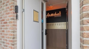 LâentrĂ©e dâun hĂŽtel, avec un mur en briques, une enseigne au nĂ©on indiquant « Amsterdam Group Stay » et une porte dotĂ©e dâune serrure de sĂ©curitĂ©.