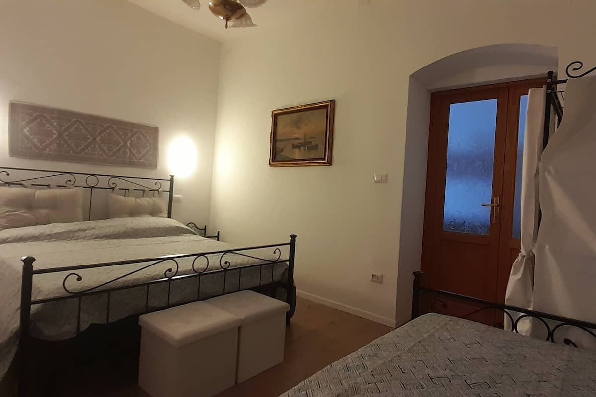 A bedroom with a metal bed frame, a bedside table, a bench, a door with a view, and a wall-mounted light.