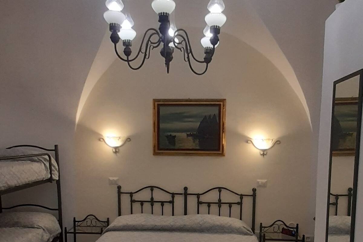 A bedroom with a large bed, a wooden bench, a chandelier, wall sconces, a painting, and a mirror.