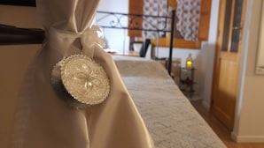 A room with a bed, a curtain, a decorative pillow, a nightstand, a lamp, and a window with curtains. - I Cappuccini B&B (Sorso)