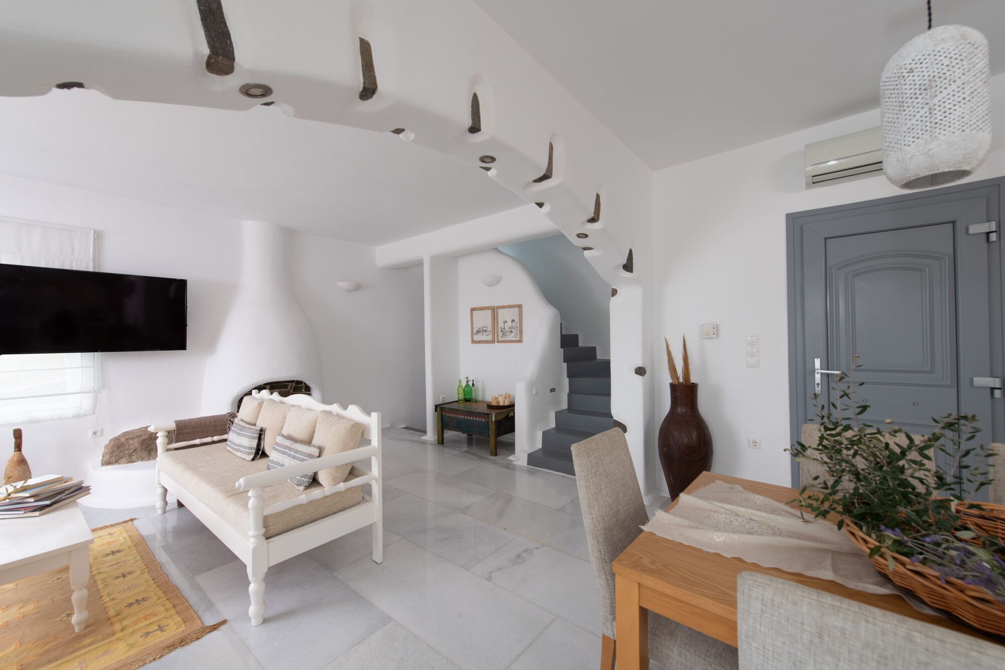 Una sala de estar moderna con un sofá blanco, una mesa de centro de madera y un televisor montado en la pared. Se ve una escalera que conduce a otro nivel y una puerta a la derecha.