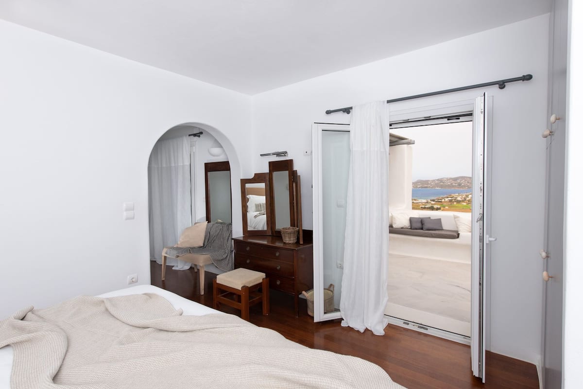 Ein Schlafzimmer mit Bett, Schminktisch, Spiegel und Meerblick durch eine offene Tür.