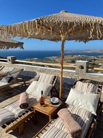 A terrace with striped lounge chairs, a table with a bottle and glasses, and a large woven umbrella.