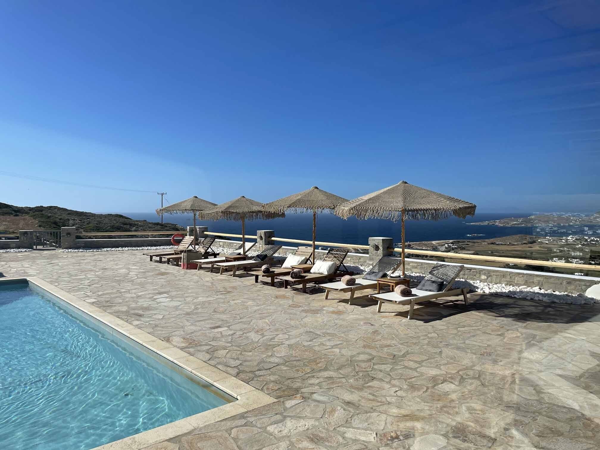 A poolside area with lounge chairs and umbrellas, a clear view of the sea, and a stone-tiled floor.