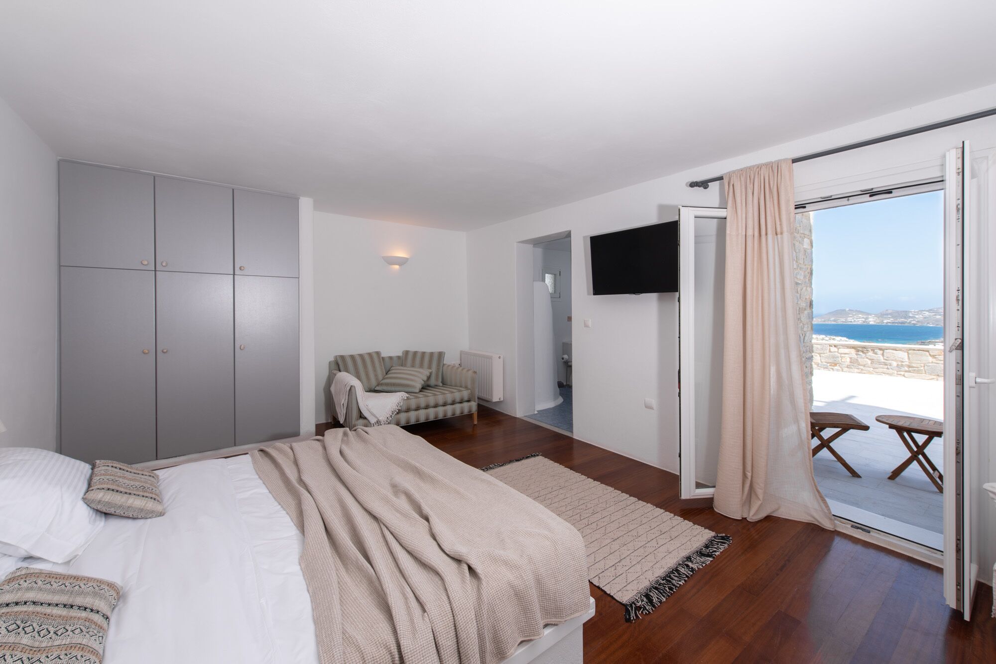Un dormitorio con cama, un armario gris, un sofá, televisor y balcón con vista al mar.