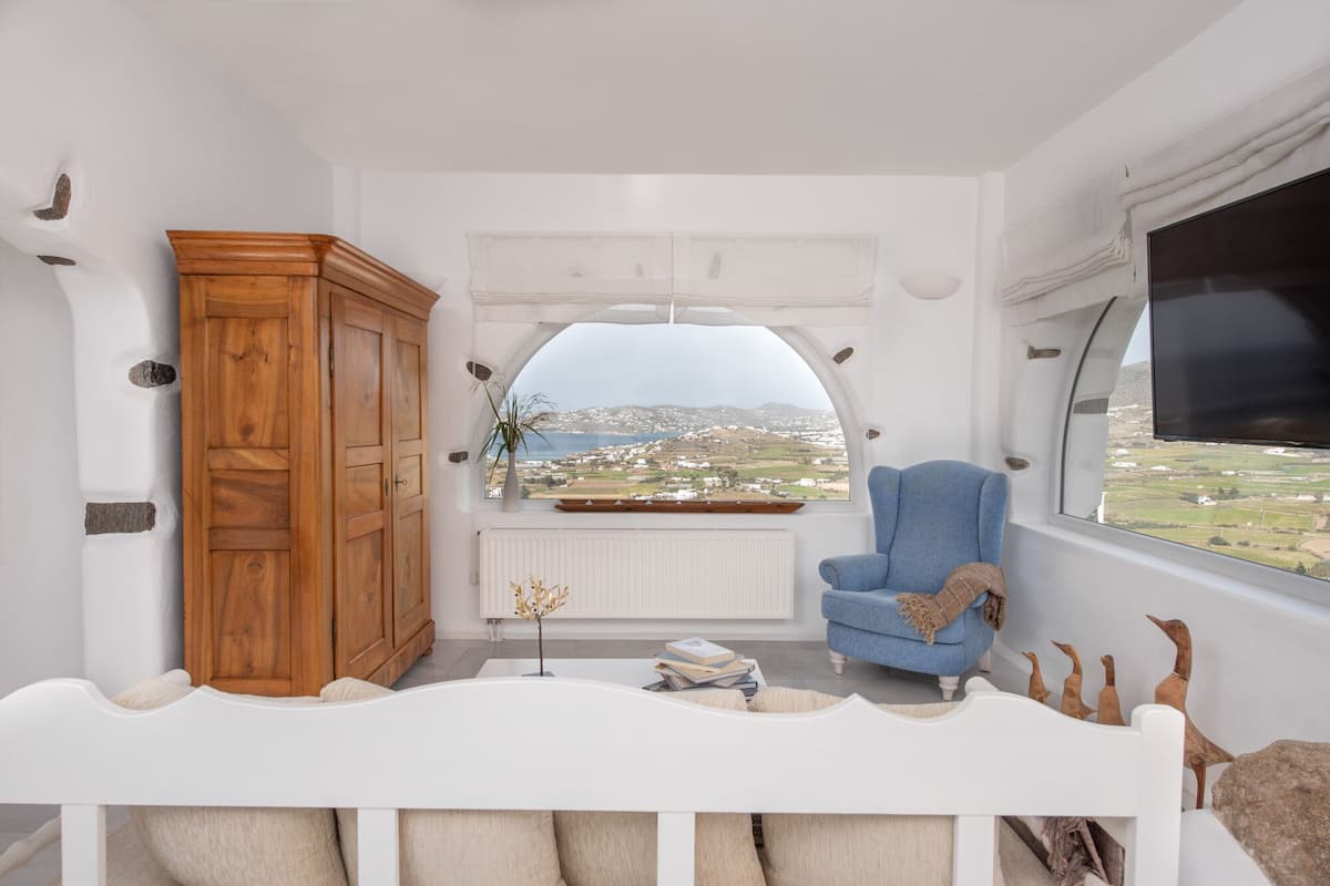 Ein gemütliches Zimmer mit einem Holzschrank, einem blauen Sessel und Blick auf eine Küstenlandschaft.