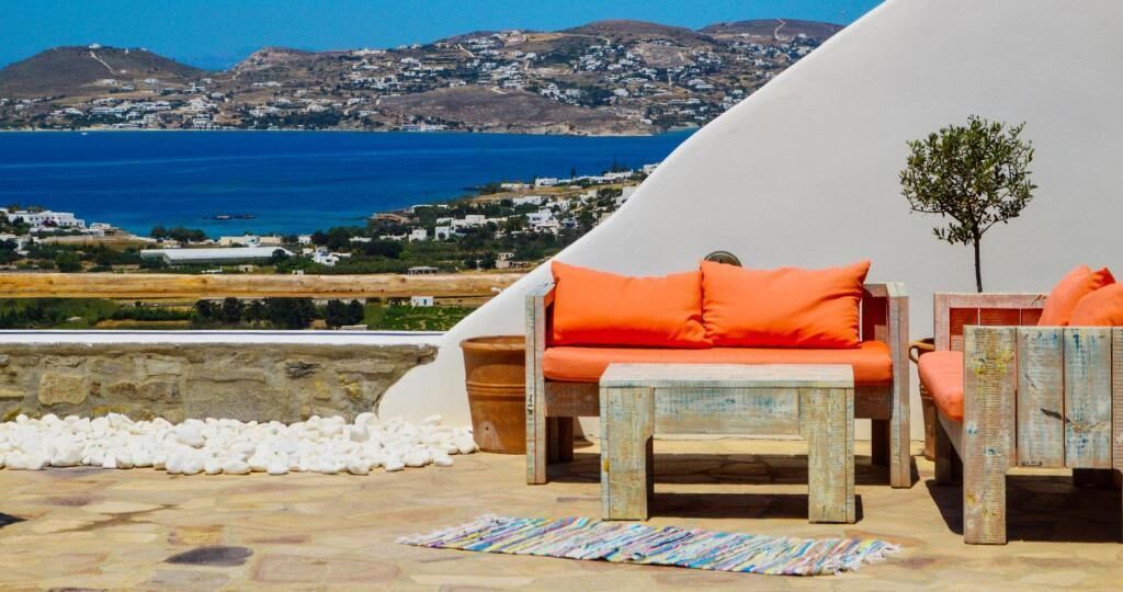 Eine Terrasse mit Blick auf das Meer und die Hügel, mit Holzmöbeln und orangefarbenen Kissen.