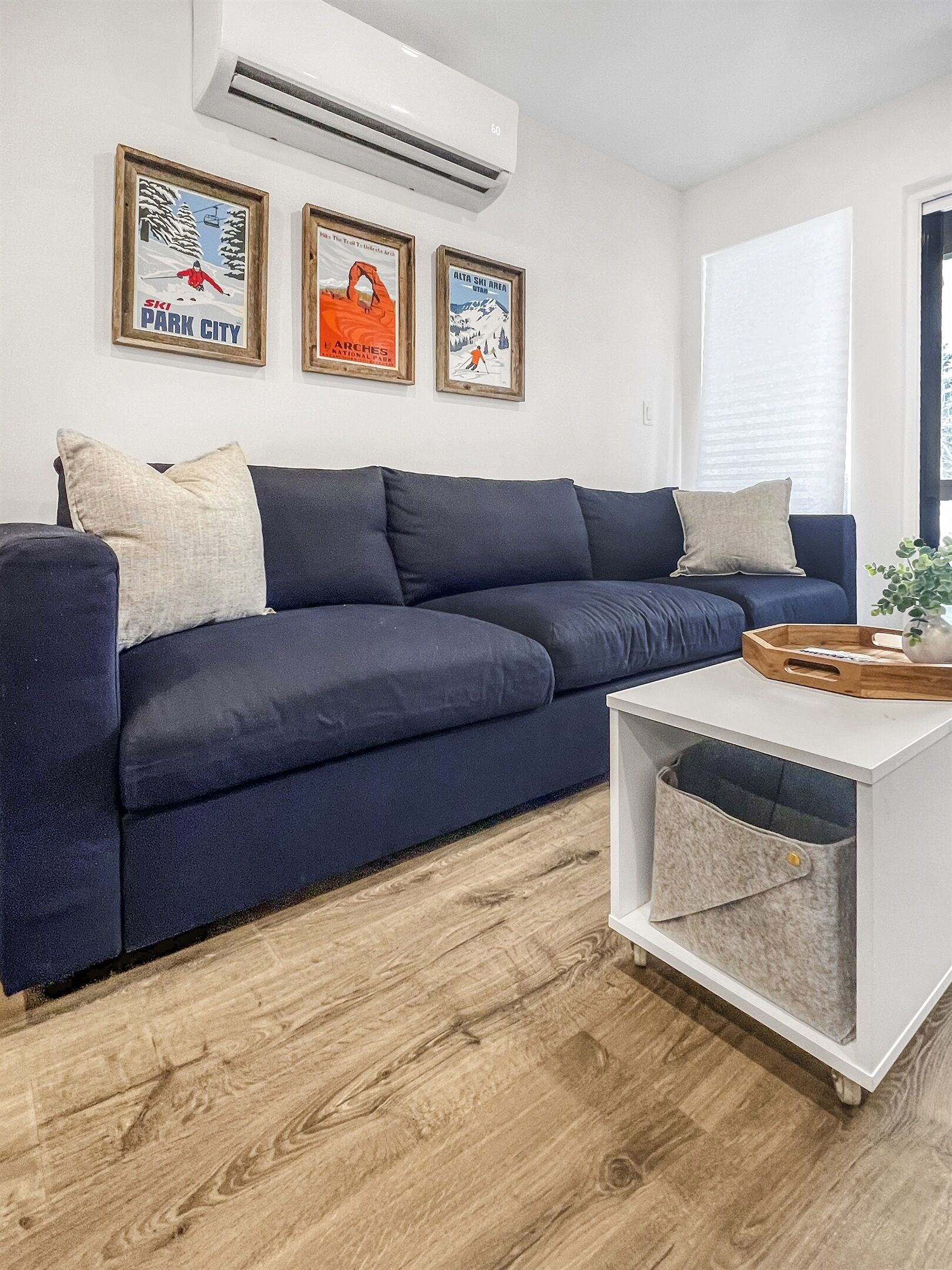 Un salon moderne avec un canapé bleu foncé, une table d’appoint blanche et un plancher en bois. Trois affiches encadrées ornent le mur, et un appareil de climatisation est installé au-dessus du canapé.